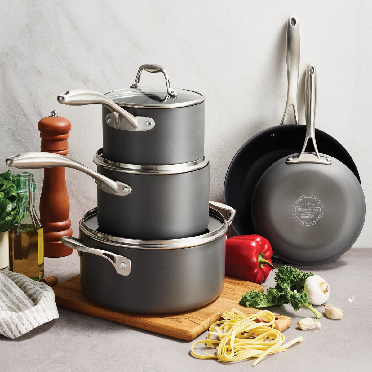 Set of gray cookware with pots, pans, and a cutting board with vegetables on a light background