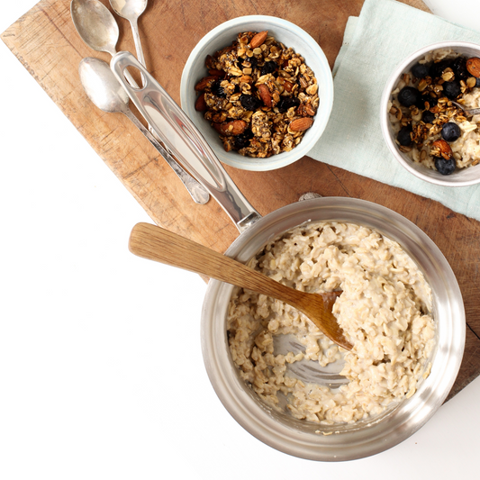 Bowl of oatmeal with a wooden spoon, surrounded by bowls of granola and blueberries on a wooden board.