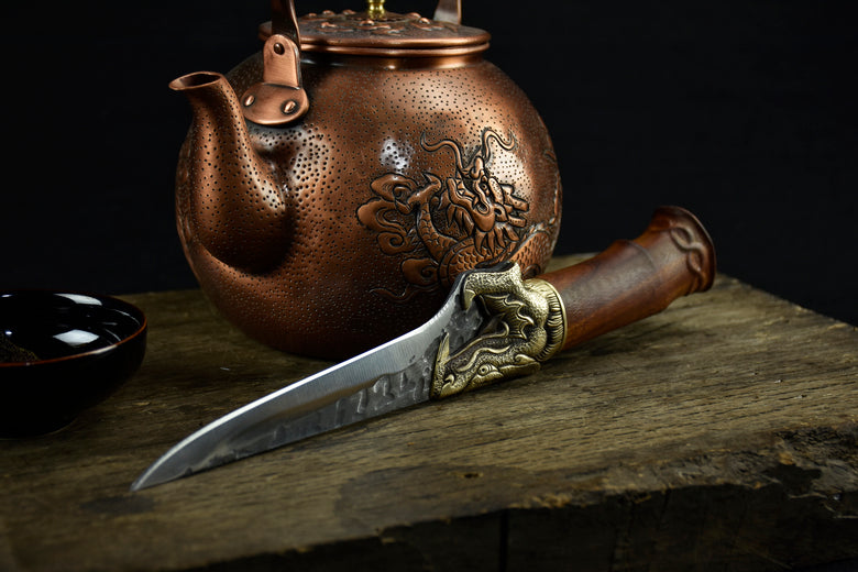 Copper teapot with embossed design and a knife with ornate handle on a wooden surface.