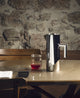 Silver pitcher and glass with red liquid on a stone table against a stone wall.
