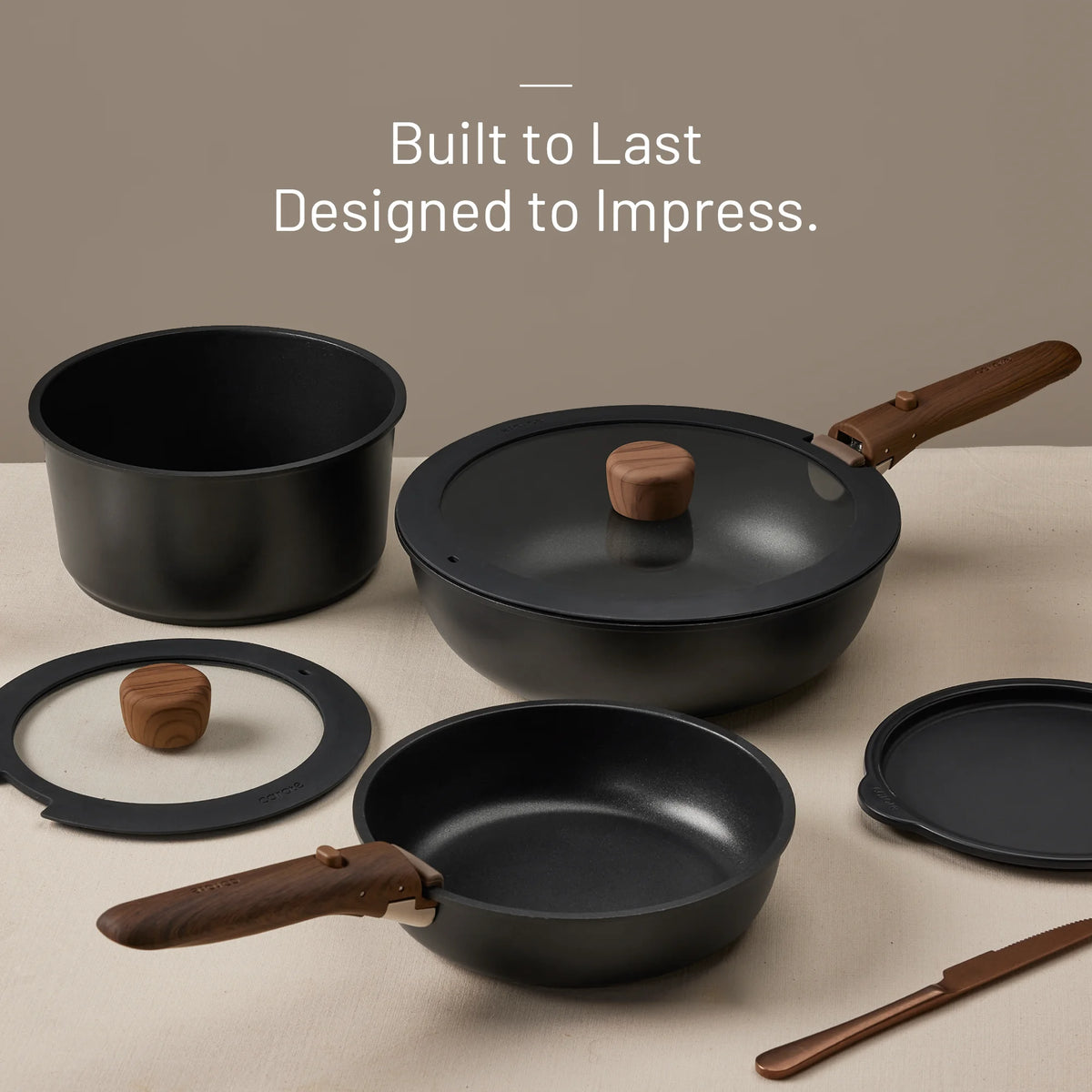 Set of black cookware with wooden handles on a beige surface, featuring text 'Built to Last Designed to Impress.'