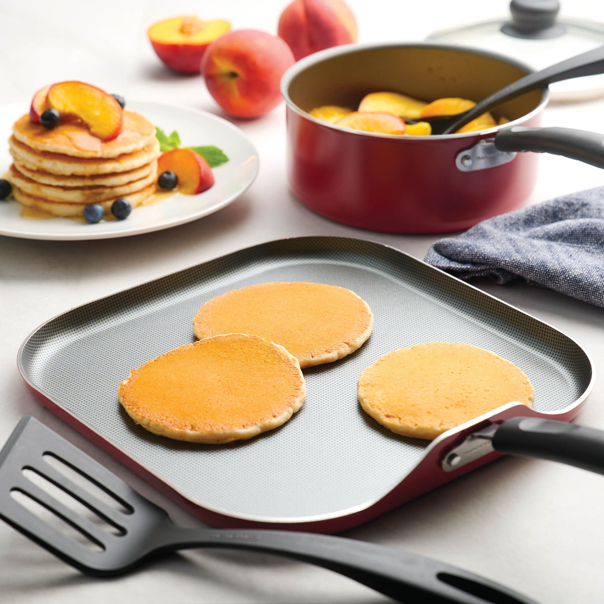 Red pot with cooked peaches and red griddle with pancakes on table