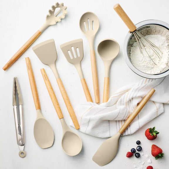 Set of kitchen utensils including spatulas, spoons, and tongs on a light surface with a towel and ingredients.