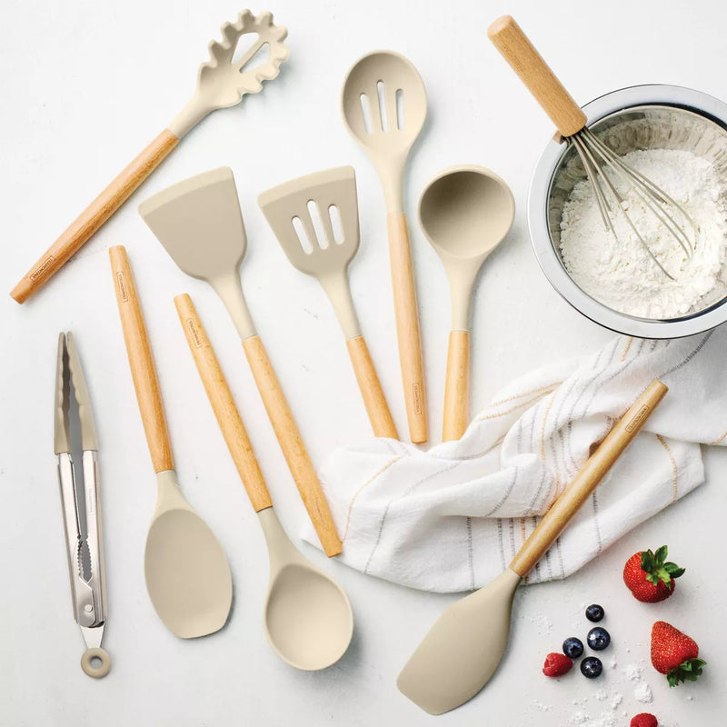 Set of kitchen utensils including spatulas, spoons, and tongs on a light surface with a towel and ingredients.