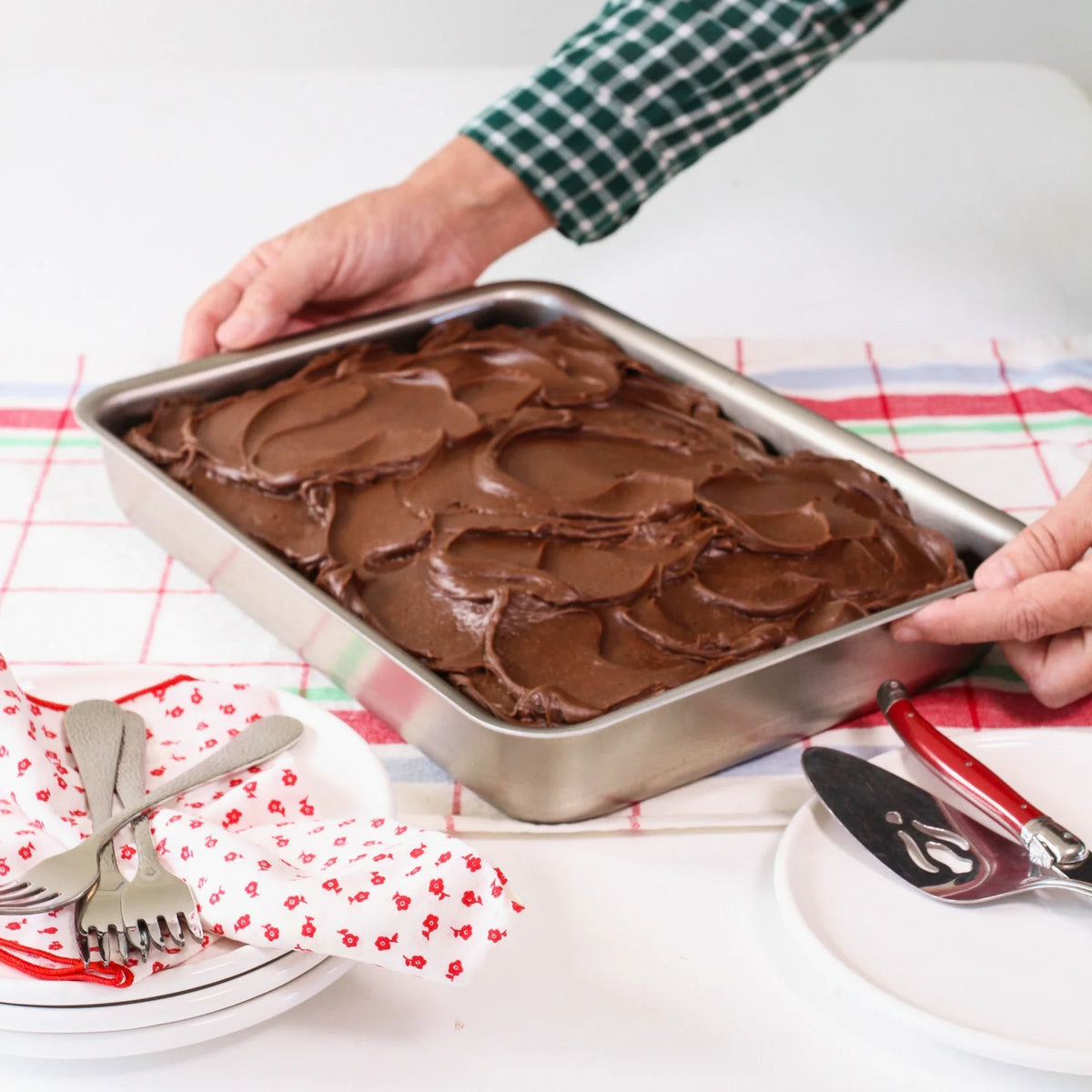 Chocolate cake in a pan on a kitchen counter with utensils and plates.