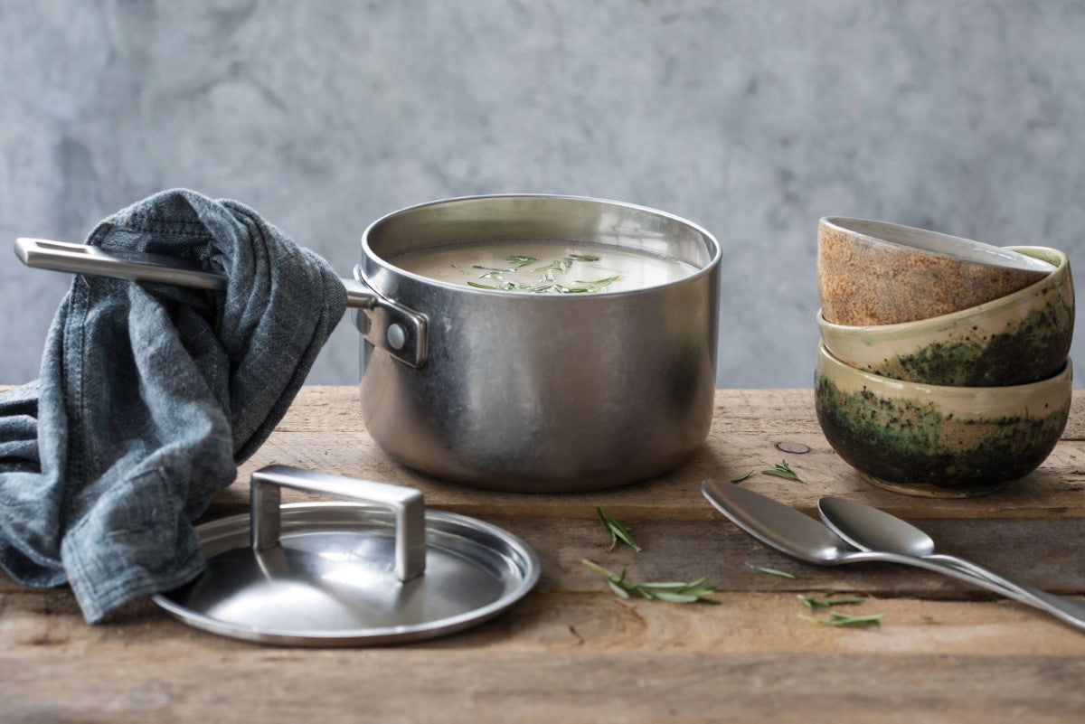 Stainless steel pot with soup, gray towel, and ceramic bowls on a wooden surface.