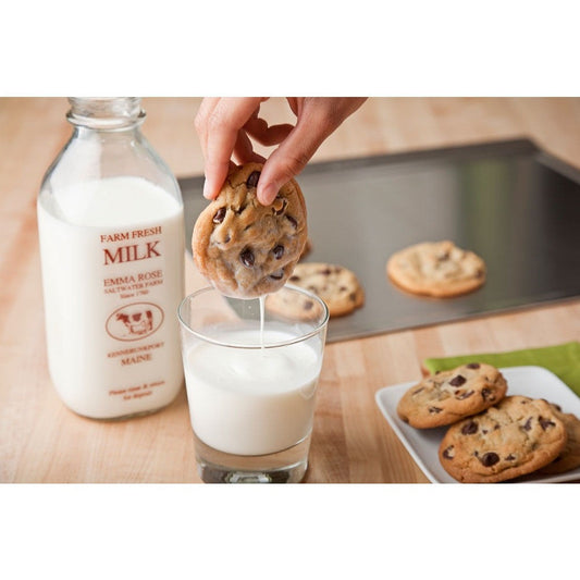 Person dipping a chocolate chip cookie into a glass of milk with a bottle of 'Farm Fresh Milk' nearby.