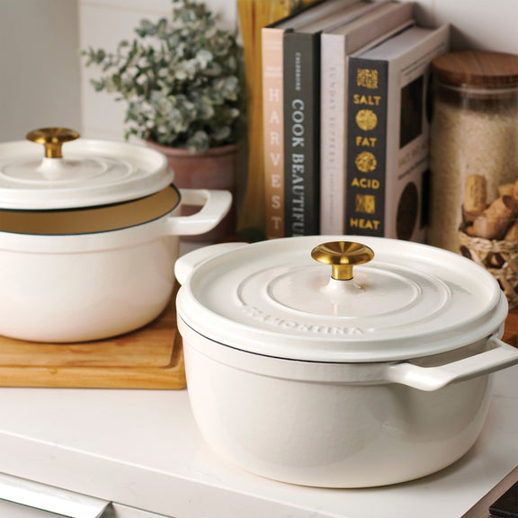 White dutch oven cookware with gold lids on a kitchen counter with cookbooks in the background