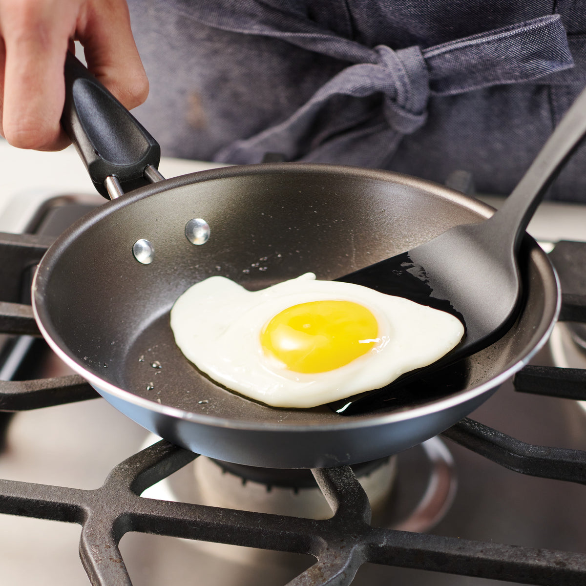 Person frying an egg in a black frying pan on a gas stove.