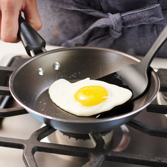 Person frying an egg in a black frying pan on a gas stove.