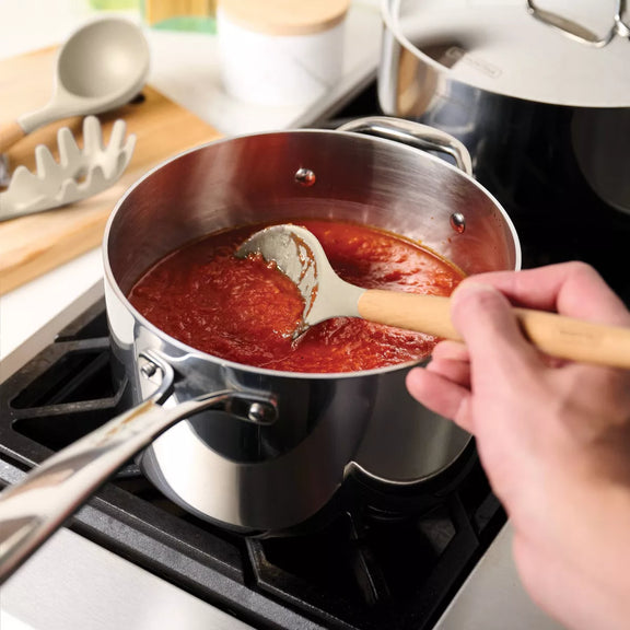 Person stirring tomato sauce in a pot on a stove with kitchen utensils in the background.