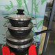 Stack of black cookware with silver handles held by a person wearing red gloves, against a blurred indoor background.