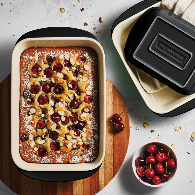Baked dish with cherries and almonds in a ceramic baking dish on a wooden board, with a container in the background.