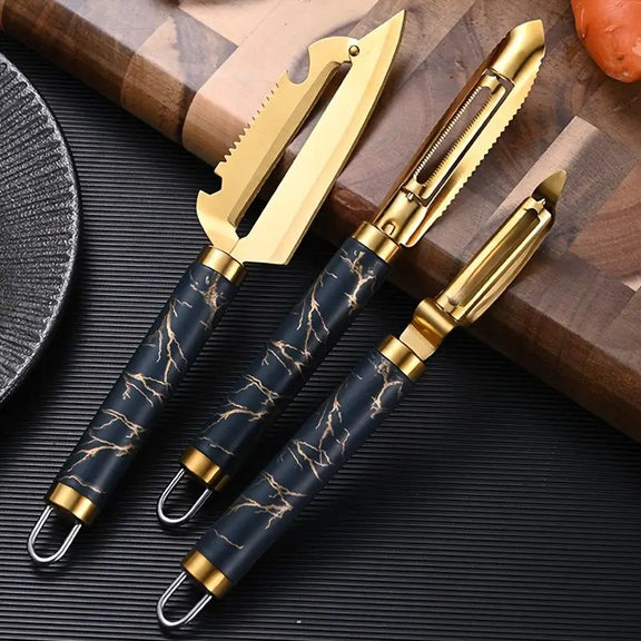 Set of three gold and black kitchen knives on a dark surface with a wooden cutting board.