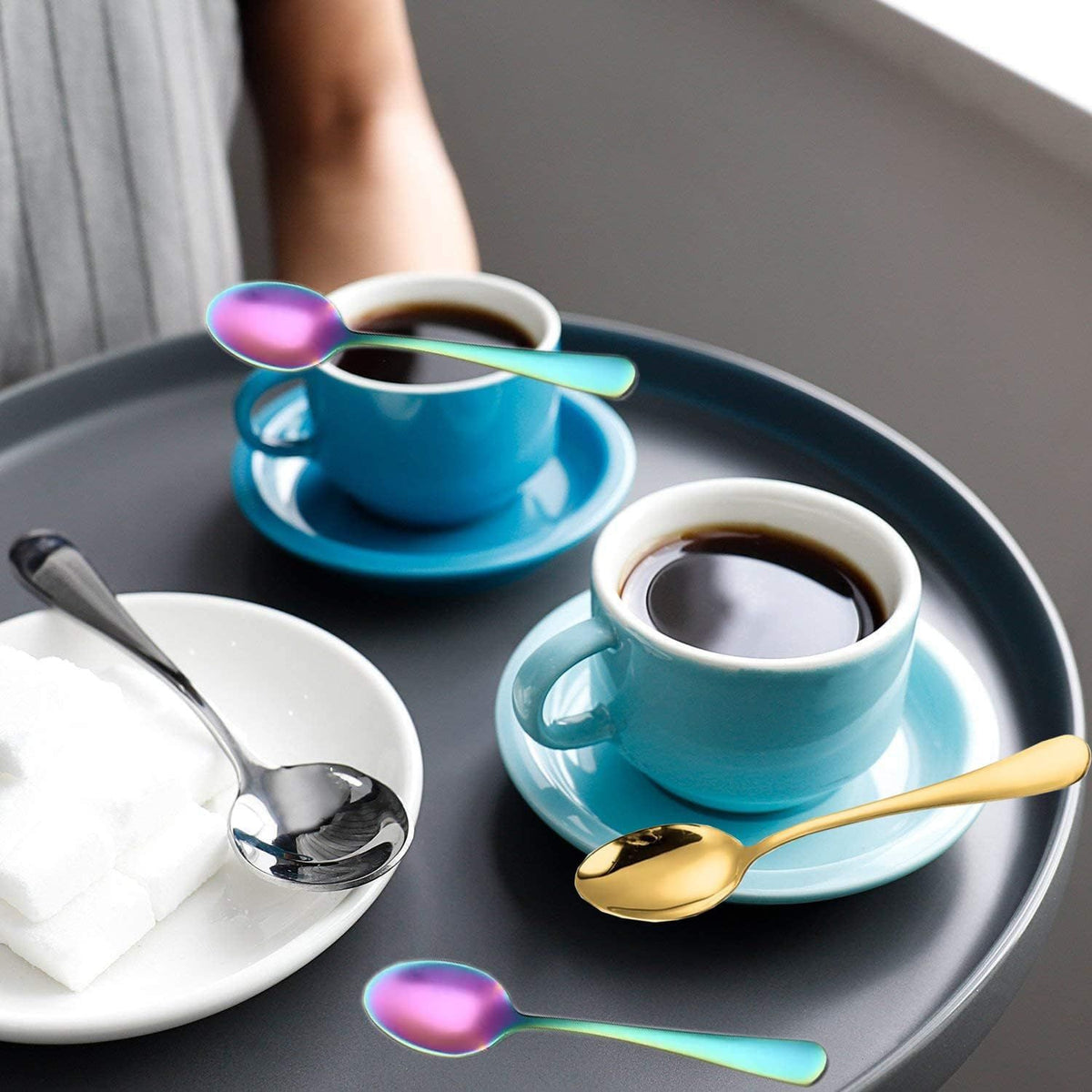 Two turquoise coffee cups with saucers on a dark tray, surrounded by colorful spoons.
