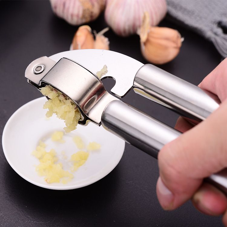 Garlic press being used to crush garlic on a dark surface with garlic bulbs in the background.