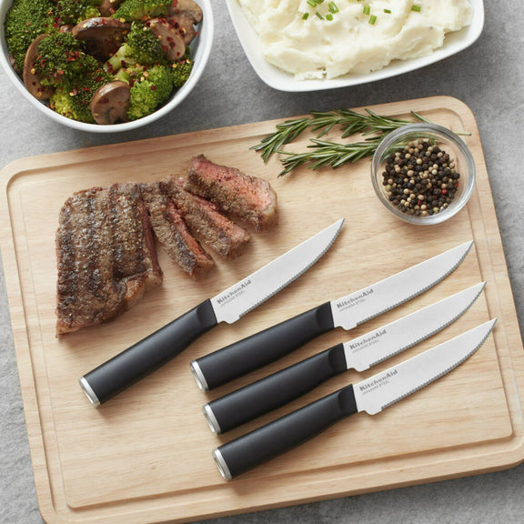Set of knives on a cutting board with sliced meat and vegetables in the background