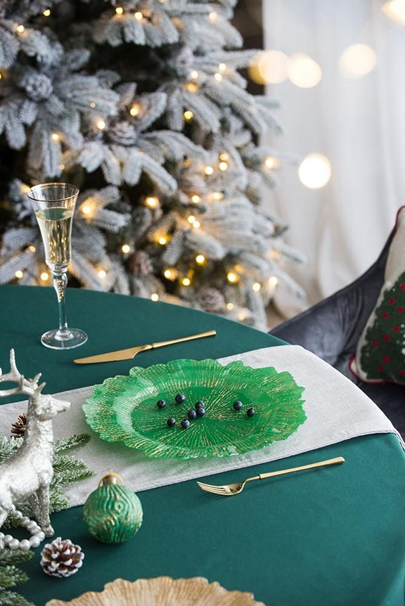 Green table setting with a decorative glass charger plate, wine glass, and gold cutlery in front of a Christmas tree.