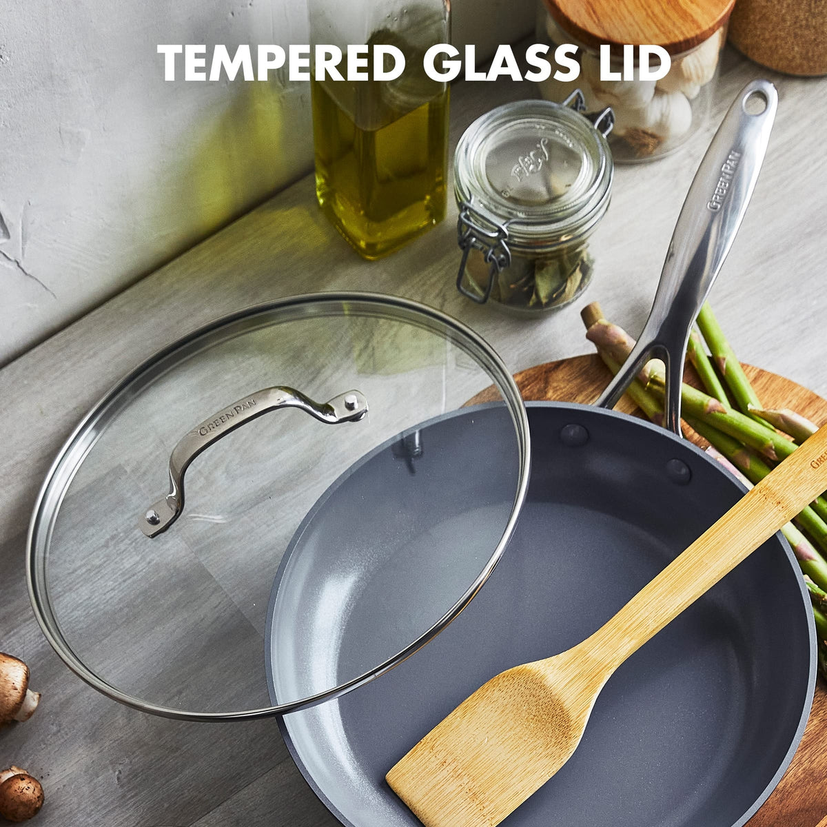 Gray Greenpan frying pan with tempered glass lid on a kitchen counter with wooden spoon and jars.