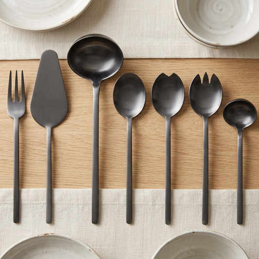 Mepra Due Ice Oro Nero seven-piece charcoal serving set on an oak table with ivory linen and ceramic dishes.
