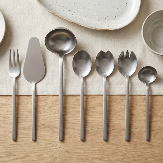 Mepra Due Ice Stainless seven-piece serving set on an oak dining table with ivory linen and ceramic dishes.