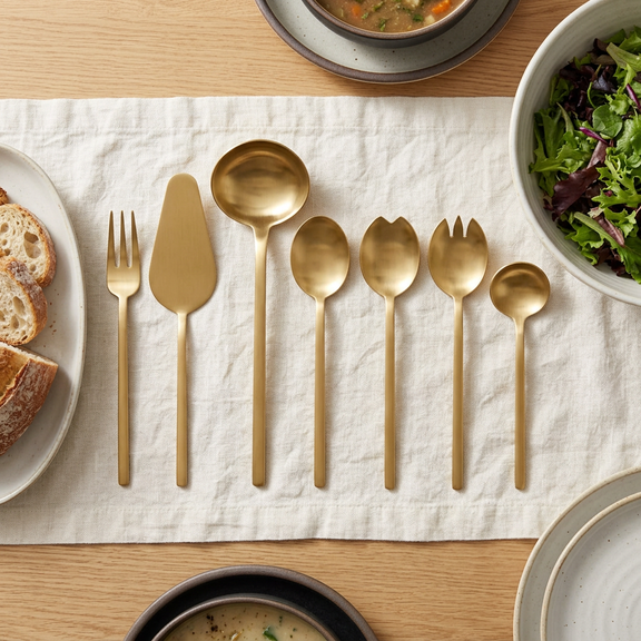 Mepra Due Ice Oro seven-piece gold serving set on ivory linen on an oak dining table.