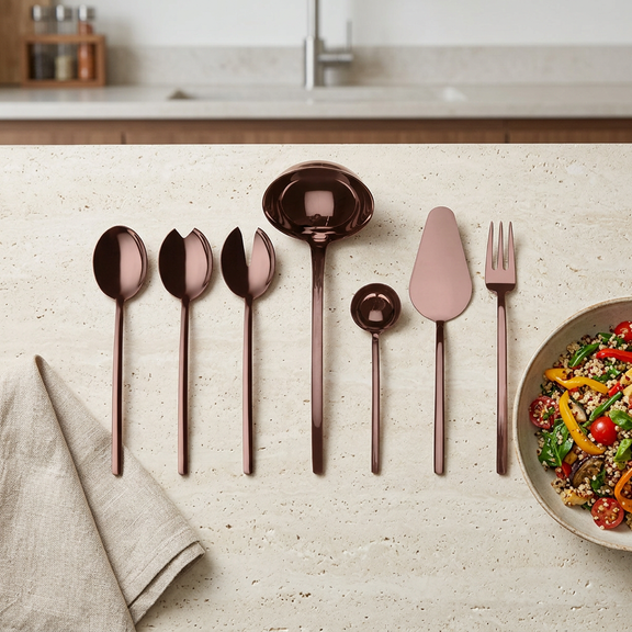 Mepra Due Bronzo seven-piece polished bronze serving set on a pale stone kitchen counter beside linen and a salad bowl.