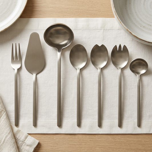 Mepra Due Ice Champagne seven-piece serving set arranged on ivory linen on an oak dining table.