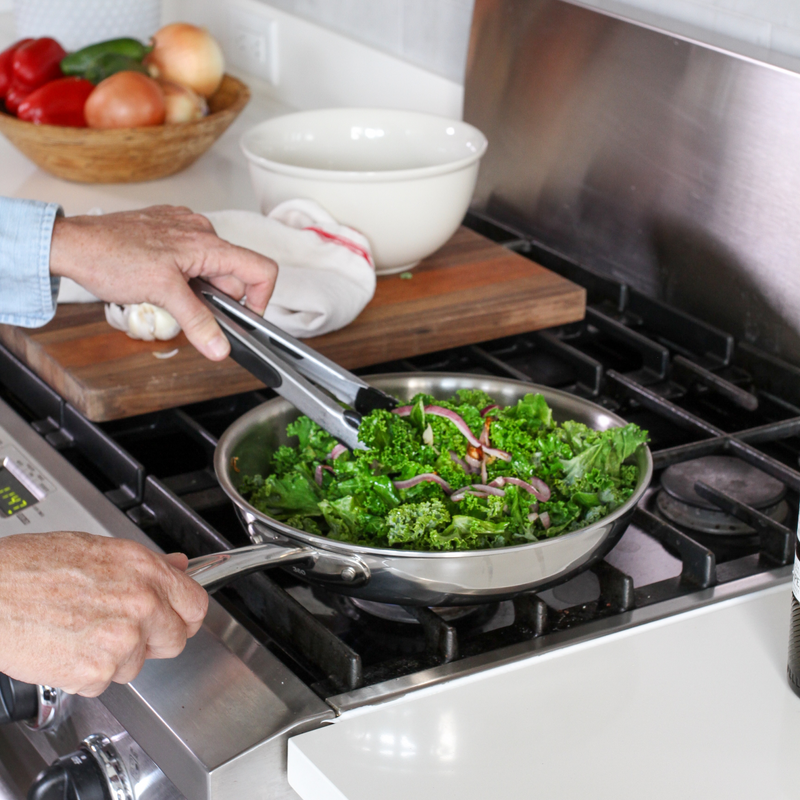 Person cooking greens in a pan on a stove with a bowl of ingredients in the background.