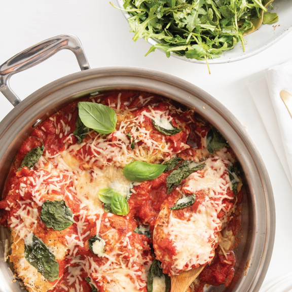 Baked dish with tomato sauce, cheese, and basil in a pan on a white surface.